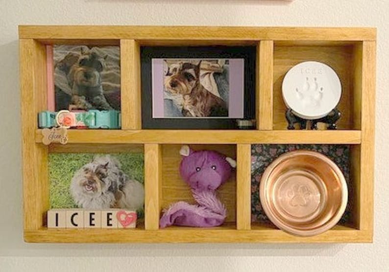 May include: A wooden pet memorial shelf with multiple compartments. It contains framed photos of dogs, a paw print keepsake, a purple plush toy, a copper dog bowl, and a dog collar. The name "ICEE" is spelled out with wooden blocks.