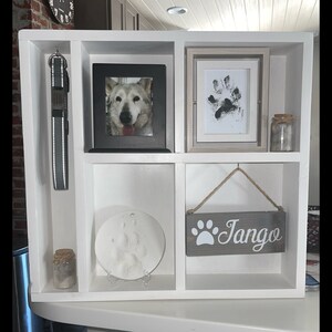 May include: A white wooden pet memorial shelf with multiple compartments. It features a framed dog photo, paw print art, a leash, and a sign that says "Tango". There are also small jars with cork lids.