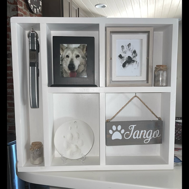 May include: A white wooden pet memorial shelf with multiple compartments. It features a framed dog photo, paw print art, a leash, and a sign that says "Tango". There are also small jars with cork lids.