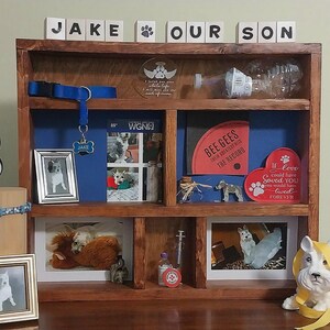 May include: A wooden display shelf with various dog-themed items. Includes framed photos, a dog collar, a dog tag that says "Jake", a heart-shaped sign, and a figurine. The shelf has the words "JAKE OUR SON" on top.