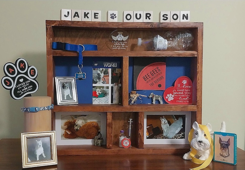 May include: A wooden display shelf with various dog-themed items. Includes framed photos, a dog collar, a dog tag that says "Jake", a heart-shaped sign, and a figurine. The shelf has the words "JAKE OUR SON" on top.