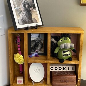 May include: A wooden pet memorial shelf with framed photos of a black poodle. The shelf contains a pink collar, a yellow flower, a paw print, a plush toy, and wooden blocks spelling "COOKIE". A small wooden box is also present.