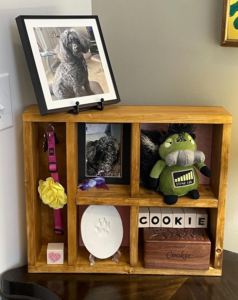 May include: A wooden pet memorial shelf with framed photos of a black poodle. The shelf contains a pink collar, a yellow flower, a paw print, a plush toy, and wooden blocks spelling "COOKIE". A small wooden box is also present.