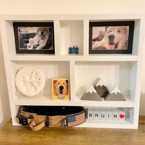 May include: A white wooden shelf unit displays framed photos of a dog, a paw print, a dog tag, a wooden block with the name "Bruin", and decorative mountain figurines. A dog collar with an American flag is also present.