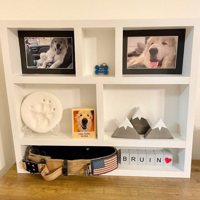 May include: A white wooden shelf unit displays framed photos of a dog, a paw print, a dog tag, a wooden block with the name "Bruin", and decorative mountain figurines. A dog collar with an American flag is also present.