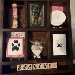 May include: A dark wooden pet memorial shelf with various items. Includes a pink dog collar, framed photos, paw print keepsakes, and a heart-shaped glass jar. The name "BLANCHE" is spelled out in wooden blocks.