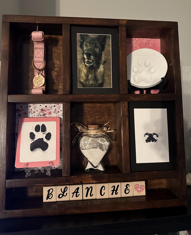 May include: A dark wooden pet memorial shelf with various items. Includes a pink dog collar, framed photos, paw print keepsakes, and a heart-shaped glass jar. The name "BLANCHE" is spelled out in wooden blocks.