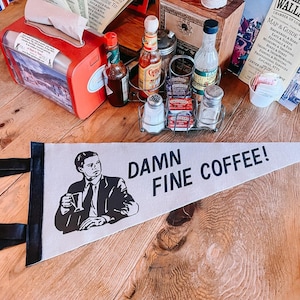 Puede incluir: Una mesa de madera con una variedad de artículos sobre ella, incluyendo un porta servilletas rojo, un pequeño banderín blanco con el texto "DAMN FINE COFFEE!" y una ilustración en blanco y negro de un hombre sosteniendo una taza de café, un pequeño termómetro rojo, una variedad de botellas y un pequeño cartel blanco con el texto "IDAHO WALLACE CHAMBER OF COMMERCE MAP & GUIDE TO: BREWERIES & BARS EATS & SLEEPS MUSEUMS & SHOPPING FESTIVALS & FUN SKIING & SNOWMOBILING BIKES & HIKES PROFESSIONAL SERVICES FISHING & HUNTING ZIPLINES, ATVS CENTRO DE PSE".