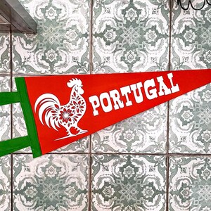 May include: A red and green pennant with a white rooster and the word "PORTUGAL" in white letters. The pennant is hanging on a wall with green and white patterned tiles.