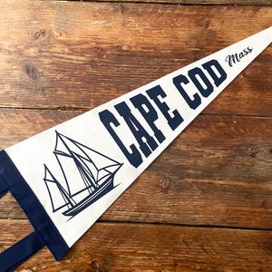 May include: A triangular felt pennant in off-white with a dark blue border and two matching ties. The design features a dark blue graphic of a sailboat and the text CAPE COD Mass in a bold, block-style font with the state abbreviation in script.