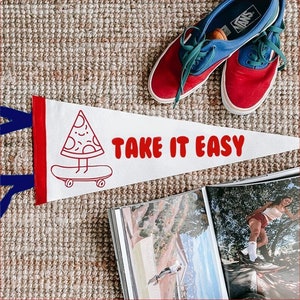Puede incluir: Un banderín blanco con ribetes rojos y azules que presenta una pizza de dibujos animados en una patineta con el texto "Take it Easy".