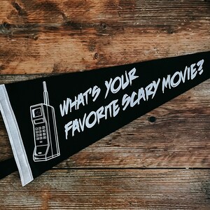 May include: Black pennant with white text that reads "What's your favourite scary movie?" and a white outline of a vintage rotary phone. The pennant is hanging on a wooden surface.