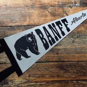 Puede incluir: Un banderín gris con la palabra "BANFF" en letras negras grandes y "Alberta" en cursiva. Una silueta de oso negro está en el banderín. El banderín tiene un borde negro y se muestra sobre una superficie de madera.