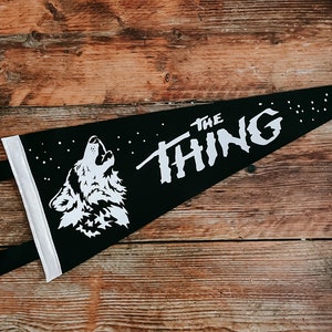 May include: Black pennant with white text that reads "THE THING" and a white silhouette of a wolf howling at the moon. The pennant is on a wooden surface.