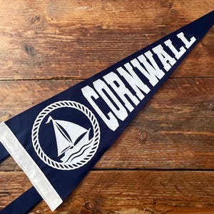 May include: A triangular navy blue felt pennant featuring the white text CORNWALL in a bold sans-serif font. Below the text is a white circular graphic of a sailboat enclosed within a rope-style border, with two white fabric ties at the base.