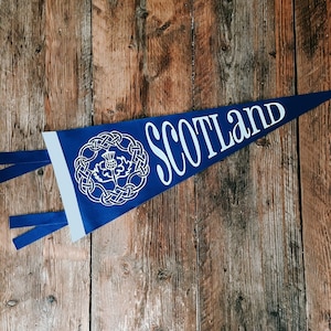 May include: Blue pennant with white text that reads "Scotland" and a white stylized thistle design. The pennant is hanging on a wooden surface.