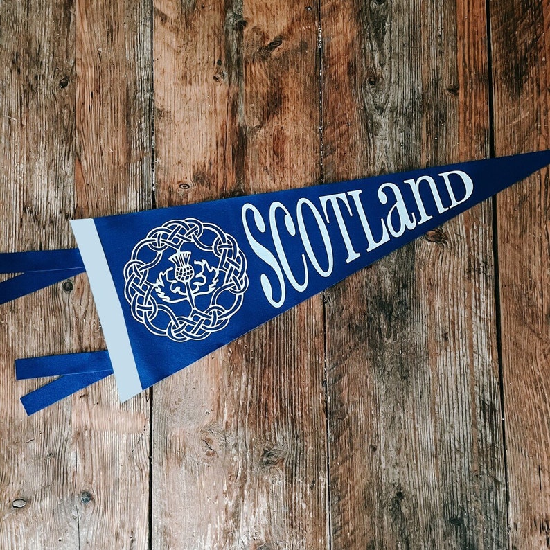 May include: Blue pennant with white text that reads "Scotland" and a white stylized thistle design. The pennant is hanging on a wooden surface.