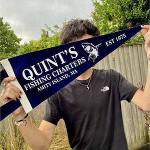 Puede incluir: Un banderín azul marino con texto blanco "QUINT'S FISHING CHARTERS AMITY ISLAND, MA EST 1975" y un gráfico de tiburón. El banderín se sostiene frente a una persona que lleva una camiseta negra.