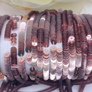 May include: A close-up of a spool of rose gold sequins. The sequins are arranged in rows and are strung together on a thin thread. The sequins are shiny and have a metallic finish.