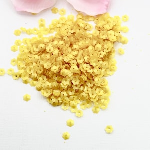 May include: A pile of small, yellow flower-shaped sequins on a white surface. A pink flower is in the background. These decorative sequins are ideal for craft projects, adding a touch of floral detail to clothing or accessories.