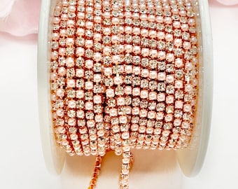 May include: A spool of rose gold rhinestone chain with alternating clear rhinestones and small pearls. The chain is on a white spool, with a portion of the chain unraveled. The background is a soft pink.