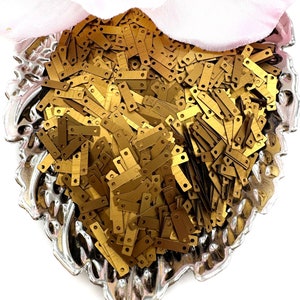 May include: A close-up view of a silver bowl filled with gold-colored metal sequins. The sequins are rectangular with two small holes at the top.