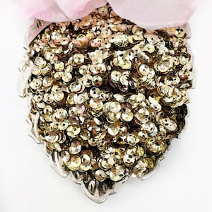 May include: A large quantity of gold sequins in a decorative silver bowl. The sequins are small and round, with a slightly iridescent finish.