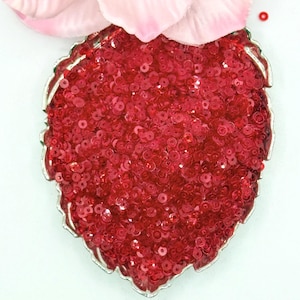 May include: A leaf-shaped brooch or patch covered in small, shiny red sequins. The object features a metallic silver-toned border with a scalloped edge. A large, pale pink artificial flower petal is positioned directly above the leaf shape.