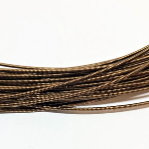 May include: Close-up of a bundle of bronze-colored, coiled metal tubing. The tubing is tightly packed together, creating a textured surface. The background is plain white, providing contrast to the metallic sheen of the coils.