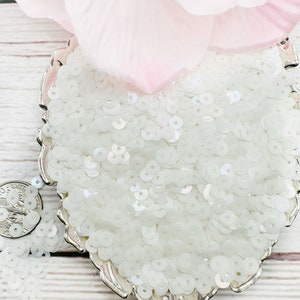 May include: A silver bowl filled with white iridescent sequins. The sequins are small and round, and they have a slight shimmer to them.
