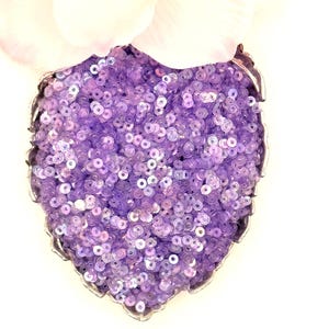 May include: A heart-shaped container filled with small, round, iridescent lavender sequins. The container has a metallic, scalloped edge that follows the heart shape, and the sequins have a reflective, shiny finish that catches the light.