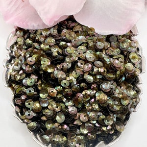 May include: A close-up of a pile of iridescent green and brown sequins. The sequins are arranged in a heart shape on a silver tray.