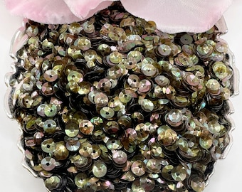 May include: A close-up of a pile of iridescent green and brown sequins. The sequins are arranged in a heart shape on a silver tray.