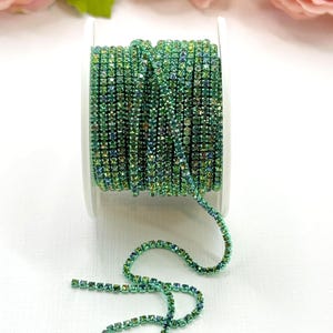 May include: A spool of green rhinestone trim. The trim is made of small, square-cut rhinestones in shades of green and teal. The trim is wound around a white spool. Pink roses are in the background.