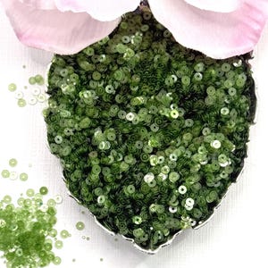 May include: A heart-shaped container filled with small, round, olive green sequins. The sequins have a reflective, metallic finish and are scattered both inside the container and on the surface next to it, with a large pale pink flower petal above.