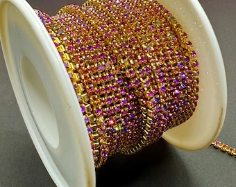 May include: A spool of gold-toned rhinestone chain with pink and purple accents. The chain is tightly wound on a white plastic spool. The rhinestones are small and closely set, creating a sparkling effect. The background is dark.