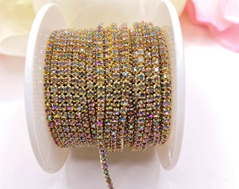 May include: A spool of gold-colored rhinestone chain. The chain is tightly wound on a white plastic spool. The rhinestones have an iridescent, rainbow-like sheen, reflecting multiple colors. The chain is partially unwound, revealing its flexible nature.