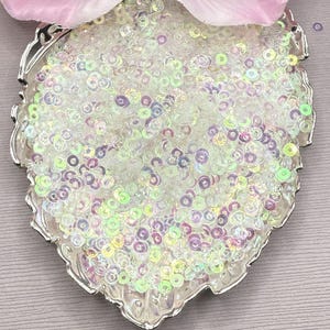 May include: A collection of small, circular, iridescent sequins with a translucent finish, displaying subtle shifts of pale green and soft purple hues. These tiny, flat, round embellishments are contained within a silver-toned, leaf-shaped dish.