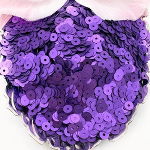 May include: A decorative, heart-shaped dish filled with vibrant purple sequins. A light pink, petal-like fabric is visible at the top of the image. The dish has a silver, scalloped edge, creating a contrast with the sequins.