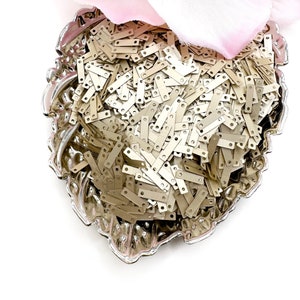 May include: A silver bowl filled with a large quantity of small, gold-colored metal sequins. The sequins are rectangular in shape with two small holes at each end.