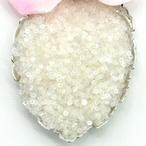 May include: A heart-shaped brooch covered in white sequins, set in a silver-toned metal frame. A pink flower is partially visible at the top. The brooch is a decorative accessory.