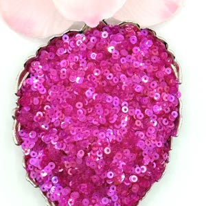 May include: A leaf-shaped pendant featuring a dense surface of vibrant hot pink sequins. The sequins are set within a metallic silver frame that follows the organic, serrated edges of the leaf shape, creating a textured and reflective appearance.