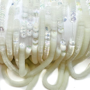 May include: A spool of iridescent white sequins, strung together in a continuous strand. The sequins are arranged in a staggered pattern, creating a shimmering effect.