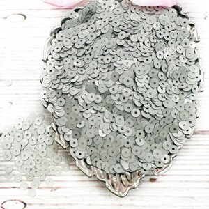 May include: A silver bowl filled with small, round, silver sequins. The sequins are scattered on a white wooden surface.