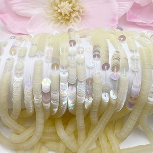 May include: A strand of iridescent white sequins with a pale yellow backing. The sequins are arranged in a row and are strung together with a thin thread. The sequins are about 1/2 inch in diameter.