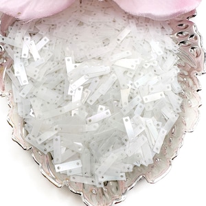 May include: A silver-colored, ornate dish filled with numerous white, rectangular sequins. A pink fabric flower is partially visible in the upper portion of the image. The sequins have small holes on each end.