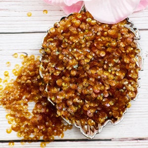 May include: A close-up of a pile of iridescent amber sequins in a silver leaf-shaped dish. The sequins are small and round, with a slightly raised center. They are scattered on a light wood surface.