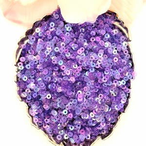 May include: A heart-shaped silver dish filled with a dense collection of small, round, purple sequins. The sequins have a reflective, iridescent finish that catches the light, creating a textured appearance within the metallic container.