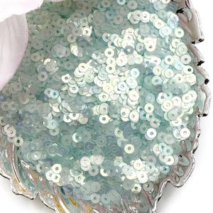May include: A collection of small, round, light blue sequins with an iridescent finish, contained within a silver-toned, leaf-shaped dish. The sequins have a flat, circular design with a central hole, reflecting light with a pearlescent sheen.