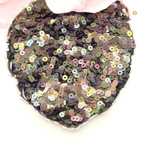 May include: A heart-shaped decorative item covered in iridescent sequins. The sequins are primarily dark brown with hints of green, purple, and gold. The item has a silver-colored scalloped edge and is partially obscured by a pink flower.